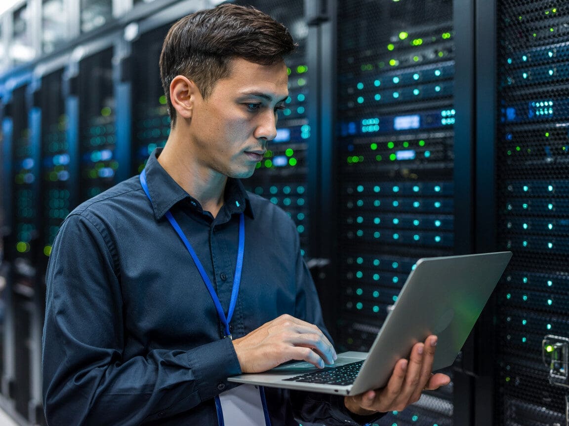 Technician using laptop in server room environment.